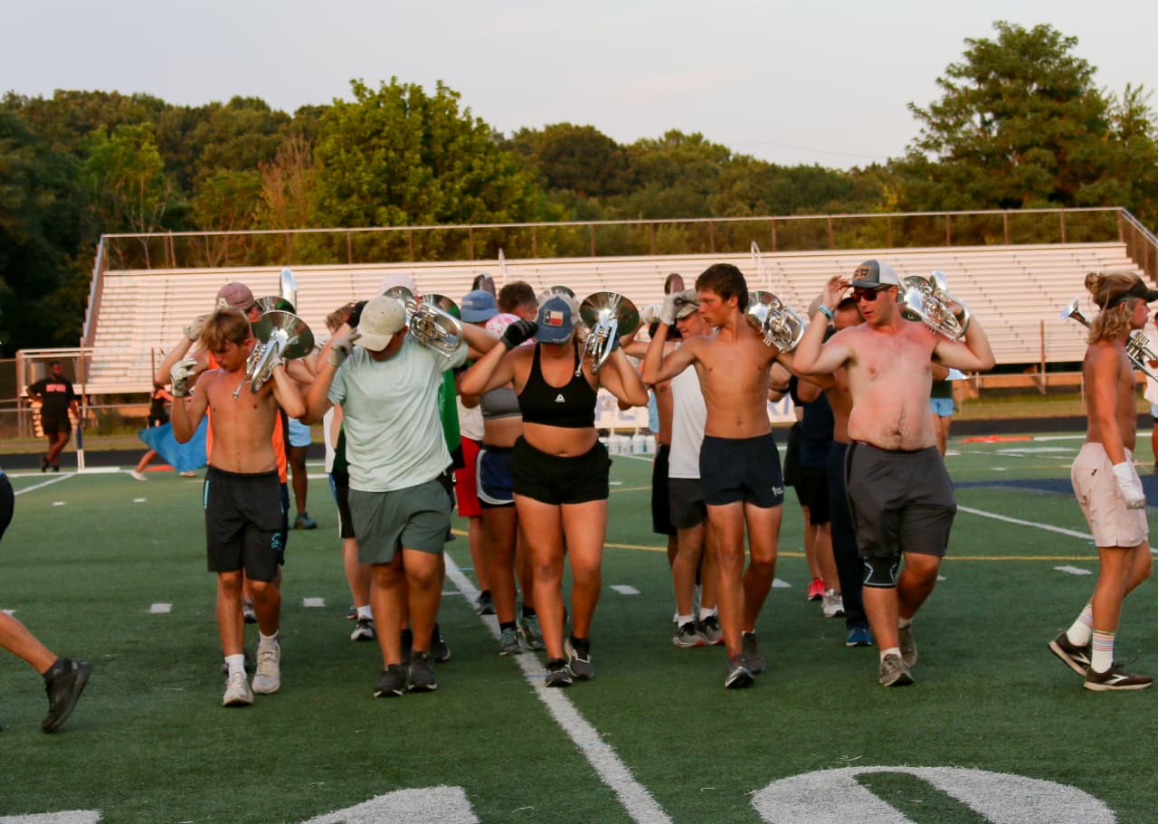Brass section carrying mellophones across the field during daytime rehearsal